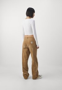 Pantalons en velours côtelé marron avec une coupe décontractée, dotés de poches arrière et d'une petite étiquette de marque. Associés à un haut à manches longues blanc.