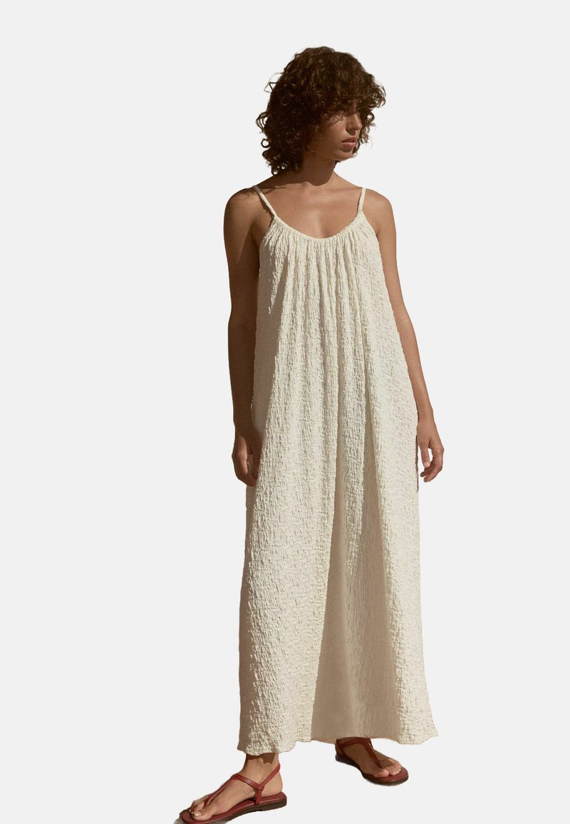 Maxikleid aus weichem, strukturiertem Creme-Stoff, mit lockerer Passform, Spaghettiträgern und gerafftem Ausschnitt. Getragen mit flachen Sandalen.
