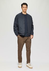 Mornarska bomberska jakna z ribastimi manšetami, temno majico, rjave chinos hlače in svetle superge. Jakna ima zadrge na žepih in gladko teksturo.