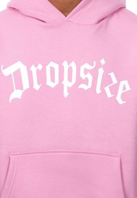 Rosa Kapuzenpullover mit einer vorderen Tasche, bedruckt mit dem auffälligen weißen Text "Dropsize" in einer verschnörkelten Schriftart. Die weiche Stoffstruktur ist sichtbar.
