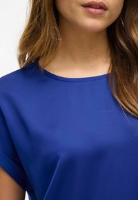 Modrý krátký rukávový top s hladkou texturou a kulatým výstřihem, mírně volný kolem ramen.