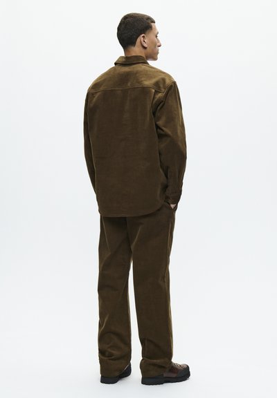 Brown corduroy shirt and matching pants; relaxed fit, visible texture, with a collared neckline and pockets. Worn with black shoes.
