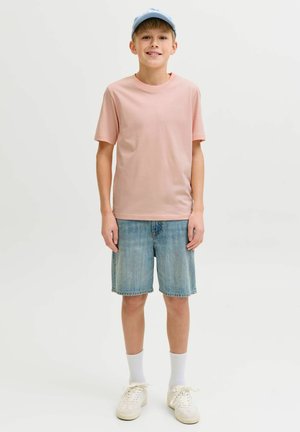 Chłopiec w jasnoniebieskiej czapce, różowej koszulce, dżinsowych szortach, białych skarpetkach i białych trampkach, stoi na tle jednolitego tła.