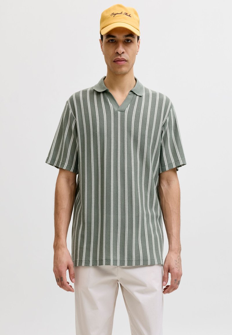 Homme portant un polo manches courtes à rayures verticales vertes et blanches, un pantalon beige, et une casquette jaune avec du texte à l'avant.