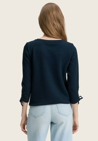 Donna con lunghi capelli castano chiaro, che indossa un top a maniche 3/4 di colore blu scuro con polsini annodati e jeans azzurri, in piedi con le spalle alla macchina fotografica.