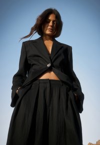 Black tailored suit featuring a cropped jacket with a single button and wide pants with pleats, paired with a simple black top underneath.