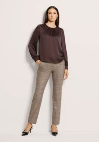 Eine Frau trägt eine dunkelbraune Satinbluse, karierte Slim-Fit-Hosen und schwarze spitze Heels mit Riemchen, während sie vor einer hellen Wand steht.