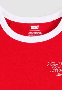 T-shirt en coton rouge avec un col rond en côte blanc. L'avant présente un texte brodé blanc indiquant "Two Toned Brand Levi's."