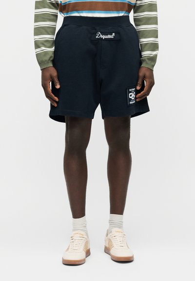 Personne portant un short noir Dsquared² avec un patch logo, une chemise à manches longues rayée, des chaussettes blanches et des baskets beige, debout droit.