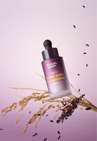 Fioletowa butelka z zakraplaczem, opisana "FIOLETOWY SERUM PDRN ZMNIEJSZAJĄCE PORY", otoczona ziarnami ryżu na gradientowym różowym tle.