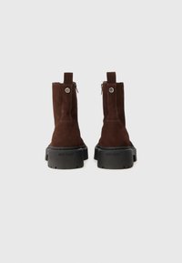 Pár tmavě hnědých semišových kotníkových bot s tlustými černými lug podrážkami, zadním zipem a značkou "Steve Madden" na patě.