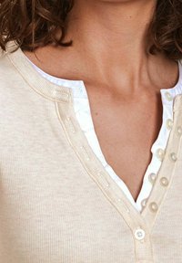 Top beige a coste con uno strato interno bianco. Presenta un collo a V e bottoni lungo la pattina, mettendo in mostra una trama liscia e un design a strati.