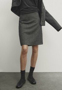 Grå tweed blyertskjol med en slät textur. Kombinerad med en svart topp, mörka strumpor och svarta skor, mot en neutral bakgrund.