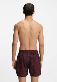 Punainen uimashortsit, joissa on tummansävyinen toistuva logokuosi, valmistettu kevyestä kankaasta, jossa on joustava vyötärö mukavuutta varten.