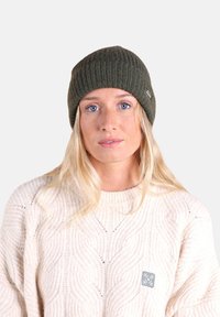 Bonnet vert olive tricoté avec une texture côtelée et une petite étiquette logo, porté avec un pull crème texturé.