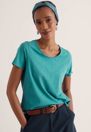 Femme portant un t-shirt bleu sarcelle, un bandeau bleu, des boucles d'oreilles bleues et un pantalon bleu marine avec une ceinture marron, debout avec une main dans la poche.