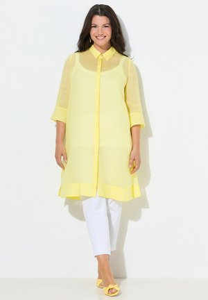 Glimlachende vrouw die een doorschijnende gele tuniek met lange mouwen draagt over witte broek en bijpassende gele openhakken, staand tegen een witte achtergrond.