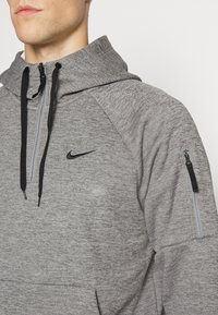 Homme portant un hoodie gris Nike avec des cordons de serrage noirs, un zip à moitié sur le devant et une poche zippée sur la manche supérieure gauche.