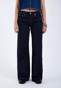 Jeans de mezclilla azul oscuro de pierna ancha con un diseño clásico de cinco bolsillos, costuras claras y un dobladillo ligeramente deshilachado en la parte recortada.