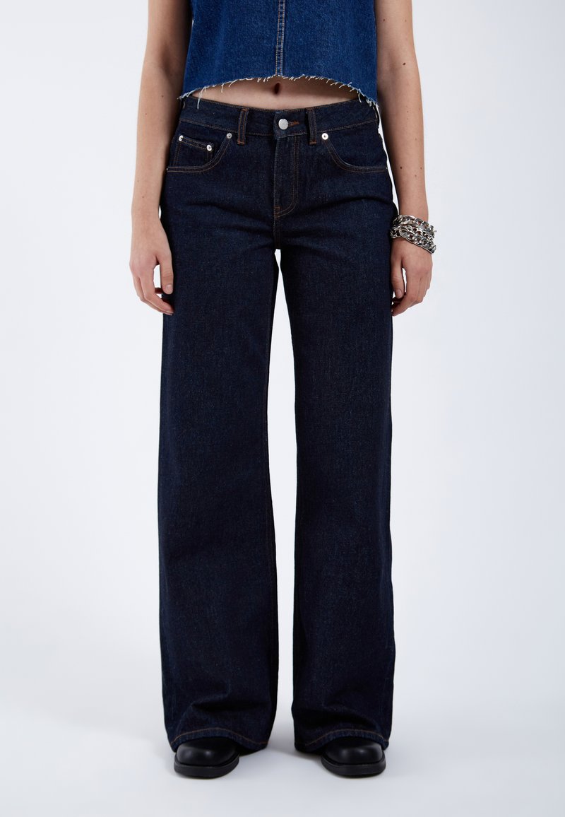 Jeans de mezclilla azul oscuro de pierna ancha con un diseño clásico de cinco bolsillos, costuras claras y un dobladillo ligeramente deshilachado en la parte recortada.