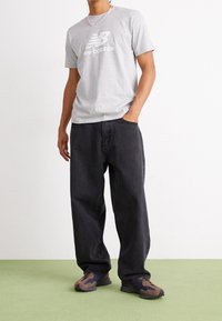 Hombre vistiendo una camiseta gris claro de New Balance, jeans negros holgados, zapatillas marrón y azul, de pie sobre un suelo verde claro contra una pared blanca.