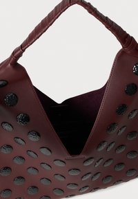 Bolsa de couro burgundy com destacados pinos redondos pretos, abertura em forma de V e pega texturizada. O interior possui um acabamento suave.