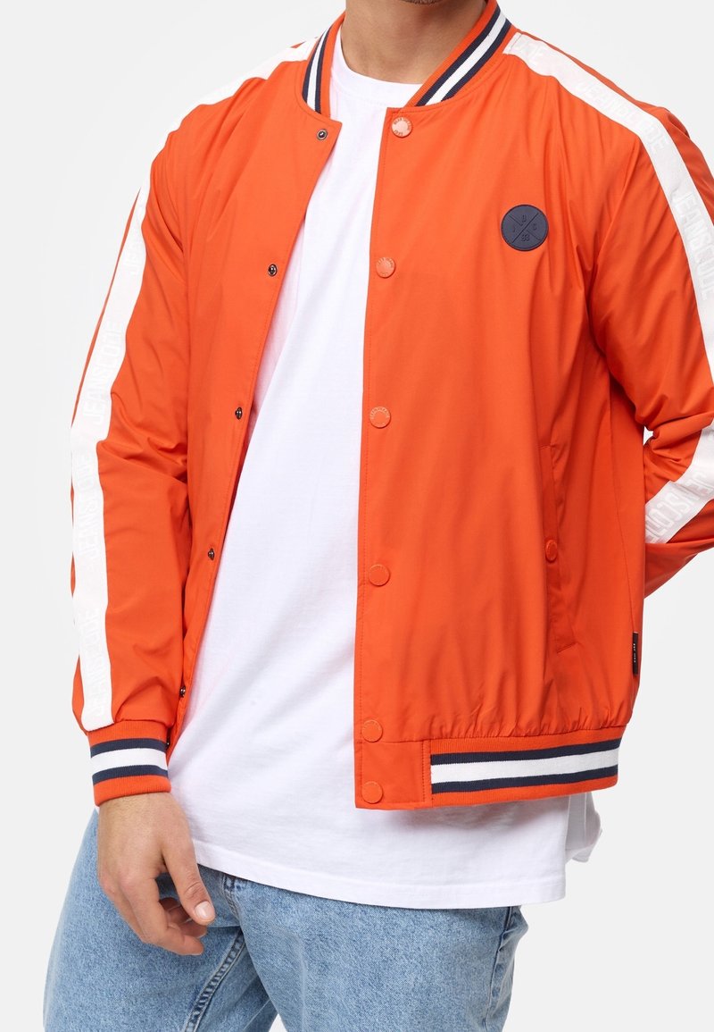 Homme portant une veste boutonnée orange vif avec des bandes blanches sur les manches, col et poignets rayés bleu marine, sur un t-shirt blanc et un jean bleu clair.