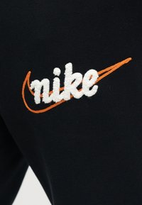 Λευκό υφασμάτινο κείμενο "nike" πάνω από ένα πορτοκαλί swoosh κεντημένο σε μαύρο υφασμάτινο φόντο.