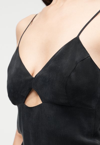 Γυναίκα που φοράει μαύρο αμάνικο φόρεμα με λεπτές τιράντες και ένα μικρό τριγωνικό άνοιγμα κάτω από το στήθος.