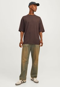 Mørkebrun oversized t-shirt parret med lysegrønne lige bukser. Modellen bærer en sort baseballkasket og beige sneakers.