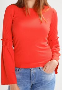 Röd ribbad långärmad topp med utsvängda ärmar som har rynkdetaljer. Bärs tillsammans med blå jeans, vilket framhäver en åtsittande silhuett.