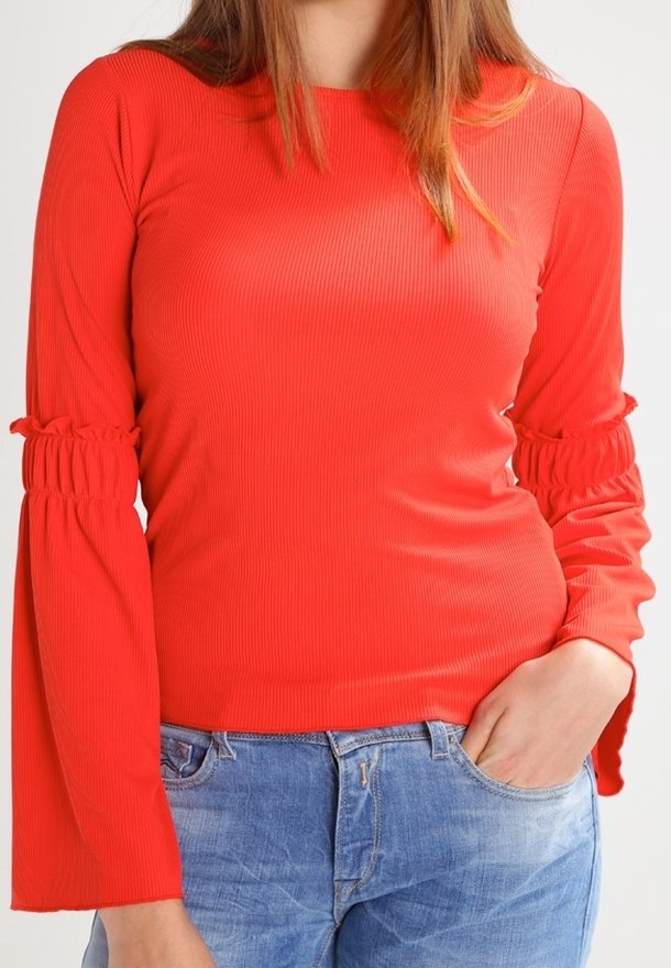 Röd ribbad långärmad topp med utsvängda ärmar som har rynkdetaljer. Bärs tillsammans med blå jeans, vilket framhäver en åtsittande silhuett.