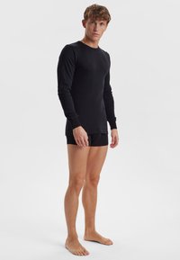 Sort langærmet skjorte lavet af glat stof, figursyet design, rund hals, båret med sorte shorts, stående barfødt mod en ensfarvet baggrund.