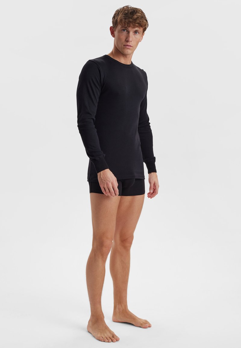 Sort langærmet skjorte lavet af glat stof, figursyet design, rund hals, båret med sorte shorts, stående barfødt mod en ensfarvet baggrund.