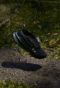 Black running shoe with textured upper, geometric accents, two white dots, and a cushioned sole, suspended above a gravel surface.