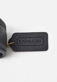 Černá kožená visačka s texturovaným povrchem, na které je uprostřed vyraženo "COACH LEATHERWARE", připevněná zlatým řetízkem.