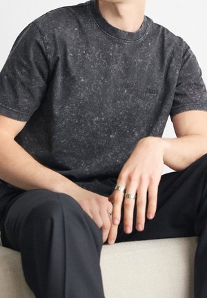 Schwarzes T-Shirt mit Marmorierung und Rundhalsausschnitt. Logo auf der Brust, weicher Stoff, lässige Passform, kombiniert mit dunklen Hosen und silbernen Ringen.