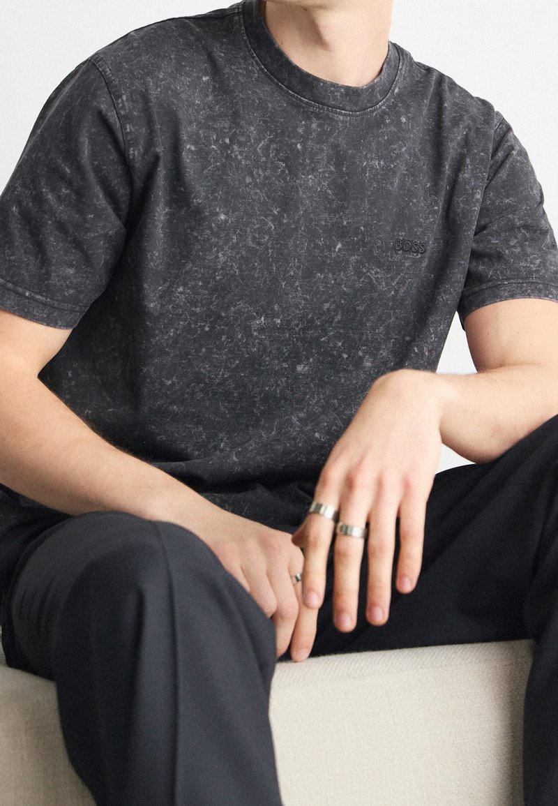 Svart T-shirt med marmor-mönster och rundad halsringning. Logotyp på bröstet, mjukt material, avslappnad passform, kombinerad med mörka byxor och silverringar.