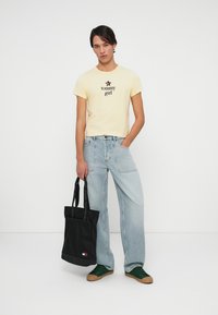 Joven vestido con una camiseta amarilla de "tommy girl", jeans azul claro, zapatillas verdes y sosteniendo una bolsa de tela negra con asas de "Tommy Jeans".