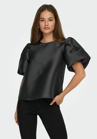 Mujer joven con cabello largo, vistiendo una blusa de satén negra con grandes mangas abullonadas y pantalones negros ajustados, posando con una mano en el bolsillo.