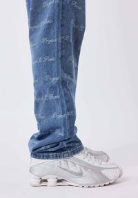 Jeans de denim con un lavado azul y un patrón de texto repetido "Forgotten" en gris claro, combinados con zapatillas Nike plateadas que presentan un diseño de suela único.