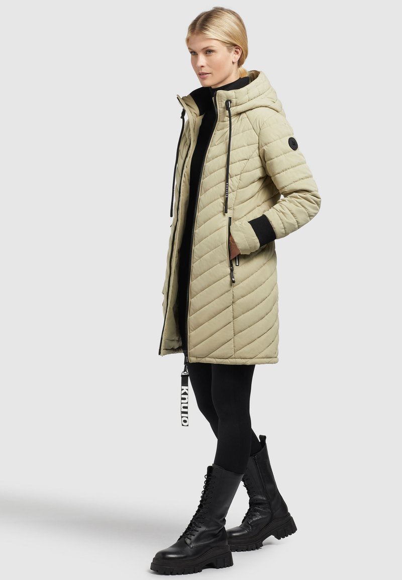 Beeindruckende, wattierte beige Daunenjacke mit Kapuze, schwarzen Akzenten, Reißverschluss auf der Vorderseite und Taschen. Kombiniert mit schwarzen Leggings und Stiefeln im Combat-Stil.