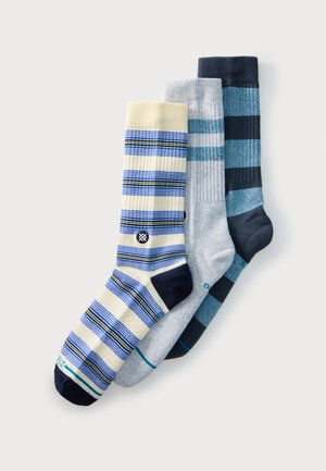 Tre par stribede sokker i beige med blå, lysegrå med teal og marineblå med lyseblå farver, arrangeret overlappende på en hvid baggrund.