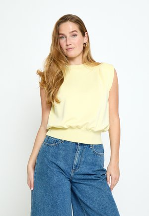 Noor naine pikkade laineliste blondide juustega, kandes varrukateta kahvatukollast pluusi ja laia säärega siniseid teksapükse, valge ühtlase taustaga.