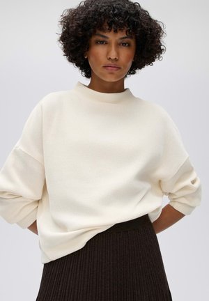 Femme aux cheveux courts et bouclés portant un pull blanc à manches longues rentré dans une jupe noire côtelée taille haute, debout devant un fond uni.