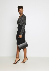 Zwarte blouse met wit abstract patroon, aansluitende zwarte midi-rok met plooien, zwarte handtas en zwarte hakken met banden. Effen achtergrond.