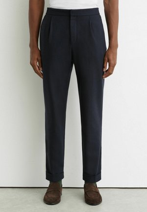 Homme portant un pantalon sur mesure bleu marine foncé avec des ourlets retroussés, des chaussettes vertes et des mocassins en daim marron, debout contre un mur blanc.