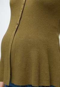 Gros plan sur un cardigan en maille côtelée vert olive avec des boutons, porté sur un jean bleu.
