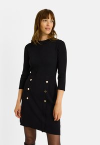 Robe noire côtelée avec un col rond, des manches trois-quarts et des boutons dorés sur le devant. Tissu lisse avec une coupe ajustée.