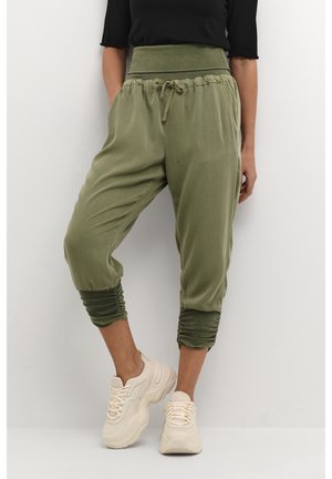 Person, der står iført olivengrønne cropped bukser med opsmøgede kanter og beige chunky sneakers, mod en ensfarvet hvid baggrund.